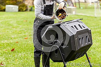 A woman is dumping a small bin of kitchen scraps into an outdoor tumbling composter in backyard garden Stock Photo