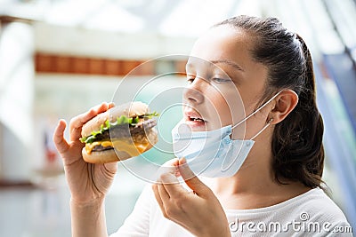 Woman Eating Fastfood Burger In Face Mask Stock Photo