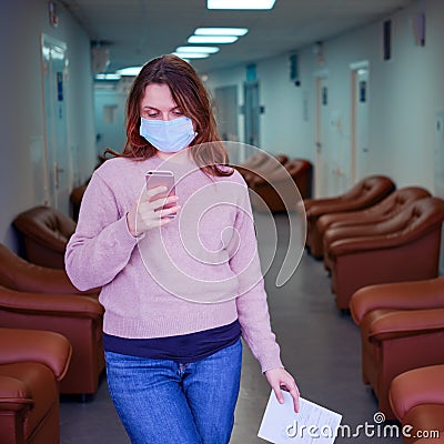 Woman in the face mask pondered in the hallway of the clinic. The patient holds a sheet of paper with a referral to the doctor Stock Photo