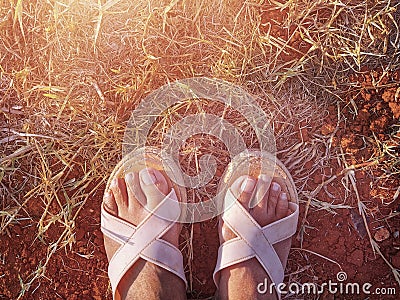 The woman feet with sandals standing on the on the dry grass field and the red clay soil Stock Photo