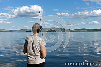 A woman fisherman stands with a fishing rod and catches fish against a blue lake and sky with clouds. Bright shot Stock Photo