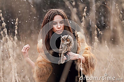 Woman in fur coat with owl on hand by first autumn snow. Beautiful brunette with long hair in nature, holding an owl. Romantic, d Stock Photo