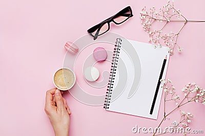 Woman hand hold cup of coffee, cake macaron, clean notebook, eyeglasses and flower on pink table from above. Female working desk. Stock Photo