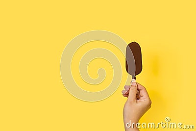 Woman hand holding a dark chocolate dipped ice lolly on a yellow background Stock Photo