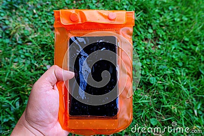 Woman hand holding the orange waterproof mobile phone case over the green grass background. PVC zip lock bag protect mobile phone Stock Photo
