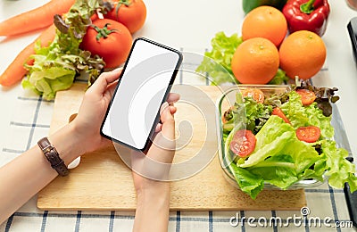 Woman hand holding a smartphone blank screen and salad bowl with tomato and various green leafy vegetables on the table. Stock Photo