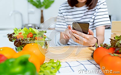 Woman hand holding a smartphone and salad bowl with tomato and various green leafy vegetables on the table at the home. Stock Photo