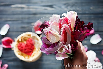 Woman hand holds bouquet of flowers tulips and red wooden heart lies in box on dark table with pink, violet petals around Stock Photo