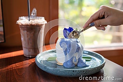 Woman hand using spoon cutting coconut cake with butterfly pea topping on ceramic dish and iced cocoa on the table Stock Photo