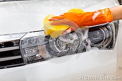 Woman hand wearing orange gloves with yellow sponge washing headlight modern car or cleaning automobile. Stock Photo