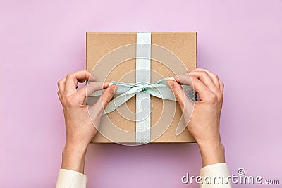 Woman hands holding craft gift box with blue bow on pastel pink background. Girl binds classic bow for present box. Top view, flat Stock Photo