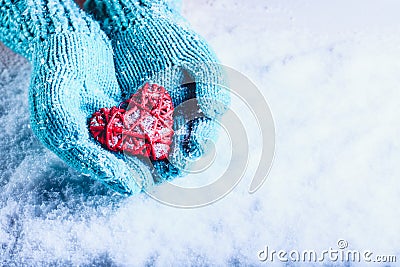 Woman hands in light teal knitted mittens are holding beautiful entwined vintage red heart in a snow. St. Valentine concept. Stock Photo