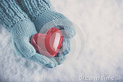 Woman hands in light teal knitted mittens are holding beautiful glossy red heart on snow background. Love, St. Valentine concept Stock Photo