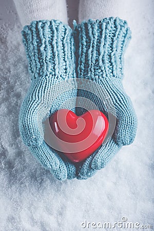 Woman hands in light teal knitted mittens are holding beautiful glossy red heart in snow background. Love, St. Valentine concept Stock Photo