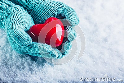 Woman hands in light teal knitted mittens are holding a beautiful glossy red heart in a snow background. St. Valentine concept. Stock Photo