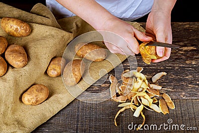 Woman hands peel potato, peelings on wooden cutting board. Three clean potatoes on plate. Stock Photo