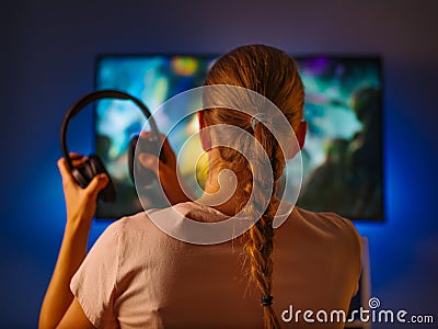 The woman has finished playing a sports video game. She holds headphones in her hands. Looks at the computer monitor. There is a Stock Photo
