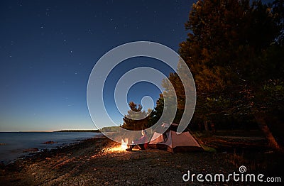 Woman having a rest at night camping near tourist tent, campfire on sea shore under starry sky Stock Photo