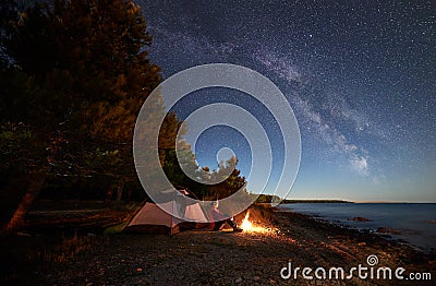 Woman having a rest at night camping near tourist tent, campfire on sea shore under starry sky Stock Photo