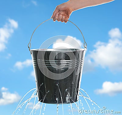 Woman holding leaky bucket with water against sky, closeup Stock Photo