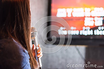 A woman singing at the karaoke bar holding a microphone in front of TV screen with lyrics. Stock Photo