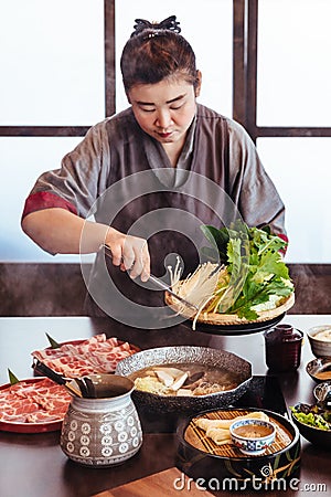 A woman holding vegetables into hot pot by tongs with Wagyu A5 beef and sliced Kurobuta in Shabu clear soup with stream Editorial Stock Photo