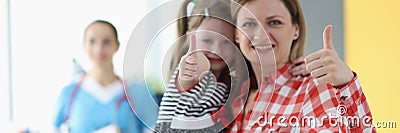 Woman holds little girl in her arms and they show thumbs up gesture on background is doctor Stock Photo