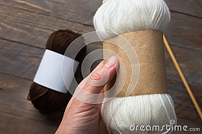 Woman holds white skein of hand`spun yarn with blank Kraft paper wrap label for mockup on rustic wooden table with needles. Stock Photo
