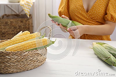 Woman husking corn cob at white wooden table, focus on basket Stock Photo