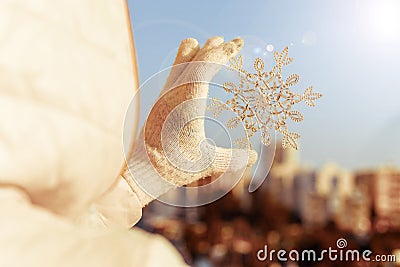 Woman in a jacket and knitted gloves holding huge sparkling snowflake on the town background in the sunset light. Winter and Chris Stock Photo