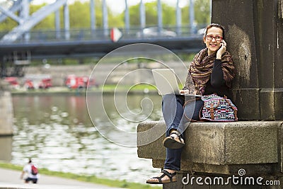 Woman with laptop sitting on the embankment of the river in the old town talking on a cell phone. Stock Photo