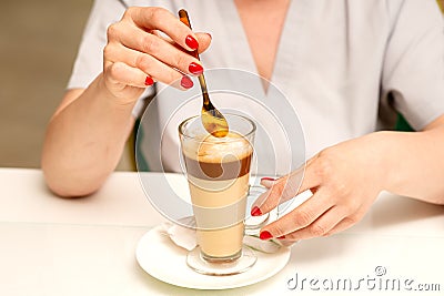 Woman with the latte. Glass mug of latte coffee on the white saucer with female hands holding teaspoon on the table in Stock Photo