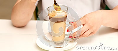 Woman with the latte. Glass mug of latte coffee on the white saucer with female hands holding teaspoon on the table in Stock Photo