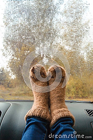 Woman legs in warm cute socks on car dashboard. Drinking warm tee on the way. Fall trip. Rain drops on windshield. Freedom travel Stock Photo