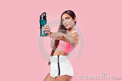 Woman with long hair muscular body in sports clothing relaxing after workout, showing botle of water on pink background. Stock Photo