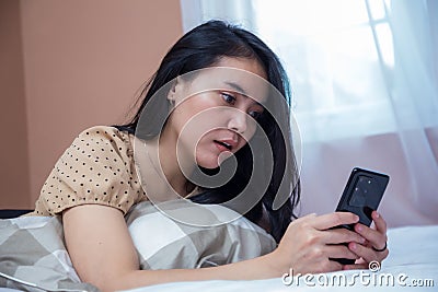 Woman lying on bed while holding cell phone with weird facial expression Stock Photo