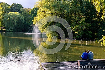 A woman and a man sitting on a wooden bench by a pond in the park Stadsparken in Lund, Sweden Editorial Stock Photo