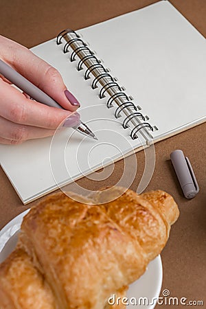 Woman with manicure writing on notebook. Freelancer working outdoors. Tasty croissant on white plate on the table Stock Photo