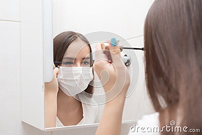 Woman in medical mask does makeup, mascaras eyes at home in the bathroom Stock Photo