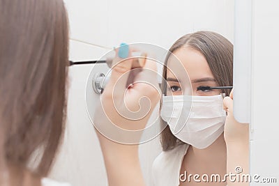 A woman in a medical mask does makeup, mascaras her eyes. Prevention and protection against diseases Stock Photo