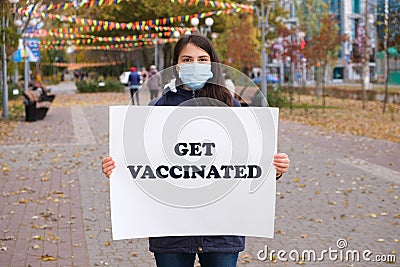 A woman in a medical mask holds a poster that says Get vaccinated Stock Photo