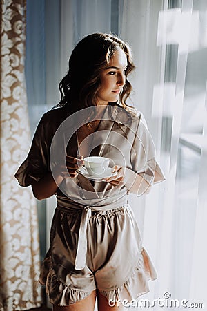 Woman in the morning. Attractive sexy woman with neat body is holding a cup with hot tea or coffee and looking at the sunrise Stock Photo
