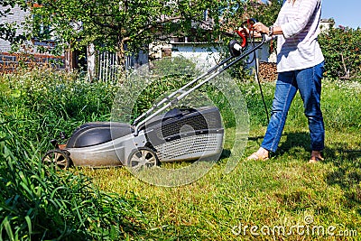 A woman mows tall green grass with an electric lawn mower in a garden plot on a summer day Stock Photo