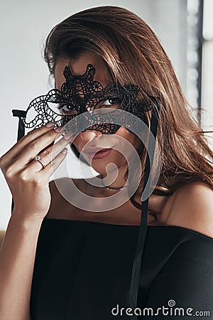 Woman is a mystery. Attractive young woman in elegant black dress wearing a black mask while standing in the room Stock Photo