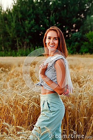 A woman in natural clothes with a string bag made of eco-mesh, fruits, apples is walking along a path in a wheat field Stock Photo