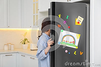 Woman opening refrigerator door with child`s drawing, notes and magnets Stock Photo