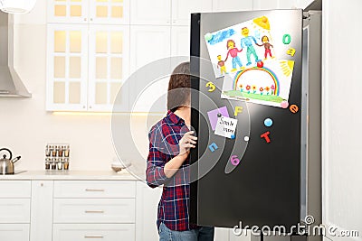 Woman opening refrigerator door with child`s drawings, notes and magnets in kitchen Stock Photo