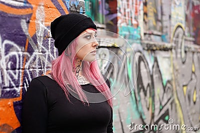 Woman with pink hair piercings and tattoos leaning against graffiti wall Stock Photo