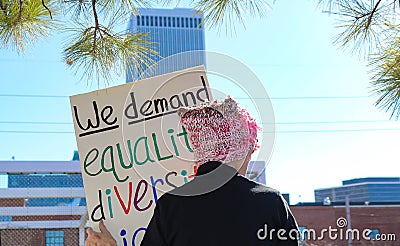 Woman n pink hat with back to camera stands against city skyline with sign We Demand Equality Diversity at Womens Editorial Stock Photo