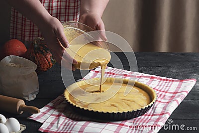 Woman pouring pumpkin pie filling from a bowl into a tray, on a black rustic table, surrounded by ingredients. Home cooking Stock Photo